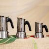 Három rozsdamentes acélból készült, fekete fogantyúval ellátott Bialetti tűzhelyi eszpresszófőző ül egymás mellett egy mintás felületen, amely a Bialetti kézi tejhabosítóval párosítva ideális otthoni krémes kávé készítéséhez.