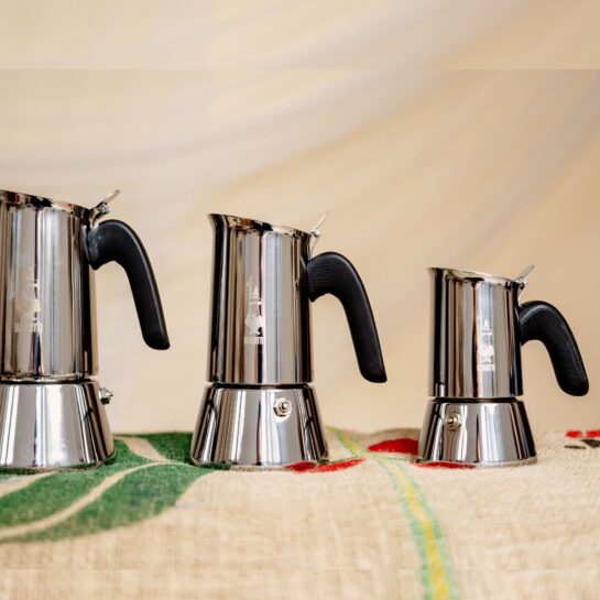 Három rozsdamentes acélból készült, fekete fogantyúval ellátott Bialetti tűzhelyi eszpresszófőző ül egymás mellett egy mintás felületen, amely a Bialetti kézi tejhabosítóval párosítva ideális otthoni krémes kávé készítéséhez.