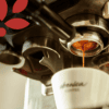 Az eszpresszó egy kávéfőző szűrőjéből egy fehér csészébe folyik, amelyen az "arabica kávé" felirat szerepel, a bal felső sarokban pedig egy piros logó egy része látható.