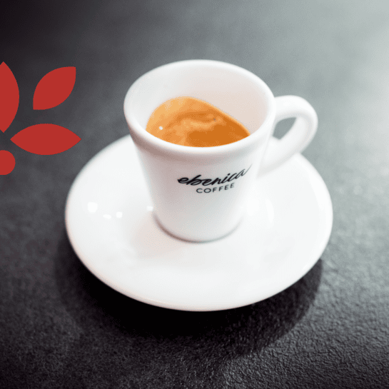 Egy fehér eszpresszócsésze, amelyen az "ebenica COFFEE" felirat olvasható, egy fehér csészealjon ül, sötét textúrájú felületen, a bal oldalon egy kis piros levélmintával.