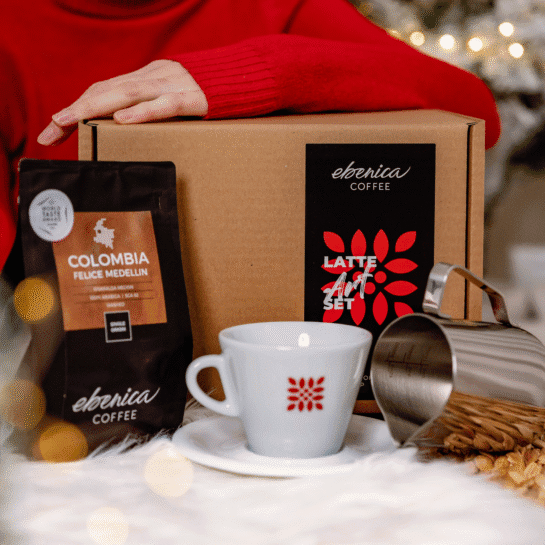 Egy kéz egy Latte Art Set dobozon pihen, mellette egy zacskó Colombia Felice Medellin kávé, egy fehér csésze csészealjjal és egy fém tejes kancsó.