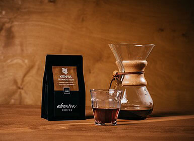 Egy zacskó kenyai kávé, egy pohár filterkávé és egy Chemex kávéfőző egy fa felületen, barna háttér előtt.