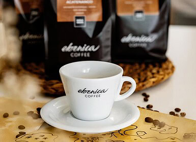 Egy fehér csésze és csészealj "ebonica COFFEE" márkajelzéssel ül az asztalon, a háttérben kávébabokkal és kávézaccokkal - tökéletes egy dupla eszpresszó elfogyasztásához.