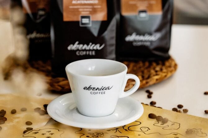 Egy fehér csésze és csészealj "ebonica COFFEE" márkajelzéssel ül az asztalon, a háttérben kávébabokkal és kávézaccokkal - tökéletes egy dupla eszpresszó elfogyasztásához.