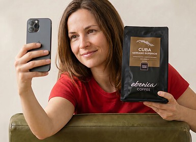 Egy piros pólós influencer egy zsák „Cuba Serrano Superior” kávét tart a kezében, és egy zöld kanapén ülve szelfit készít a telefonjával.
