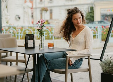 Egy nő ül egy szabadtéri kávézó asztalánál egy itallal, egy csomag kávéval és egy vázában lévő virággal – oldalra pillant, és megsimogatja a haját, mintha a közelmúltbeli influencerekkel való médiakapcsolatairól gondolkodna.