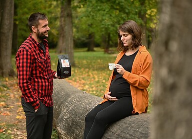 Egy nő egy kidőlt fatörzsön ül, kezében egy csészével, és simogatja terhes hasát. Mellette egy piros kockás ingben álló férfi áll egy zacskó kávéval a kezében. Ez a hangulatos szabadtéri jelenet tökéletesen alkalmas influencer-marketingre vagy médiakampányokra.