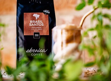 Egy zacskó Ebonica Brasil Santos kávé áll egy fémpohár és egy szívószál mögött, az előtérben pedig zöld növények láthatók – tökéletes inspiráció a következő frappé-receptedhez.