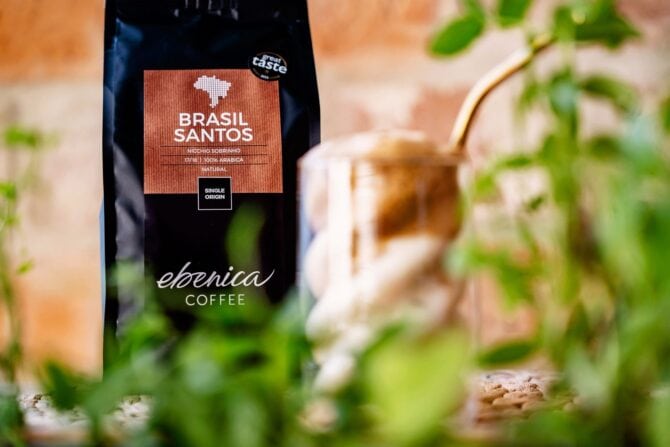 Egy zacskó Ebonica Brasil Santos kávé áll egy fémpohár és egy szívószál mögött, az előtérben pedig zöld növények láthatók – tökéletes inspiráció a következő frappé-receptedhez.
