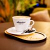 Egy fehér kávéscsésze, amelyen az „ebonica COFFEE” felirat látható, egy csészealjon áll, mellette egy kanál és egy kis keksz, mindez egy „Muse.” feliratú fa tálcán.