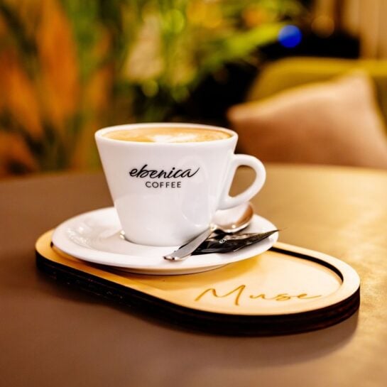 Egy fehér kávéscsésze, amelyen az „ebonica COFFEE” felirat látható, egy csészealjon áll, mellette egy kanál és egy kis keksz, mindez egy „Muse.” feliratú fa tálcán.