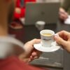 Egy személy egy kis fehér kávéscsészét tartja a kezében, amelyen az „Ebenica Coffee” felirat látható; egy asztalnál ül, mellette egy laptop és egy jegyzetfüzet, a háttérben pedig más emberek is láthatók.